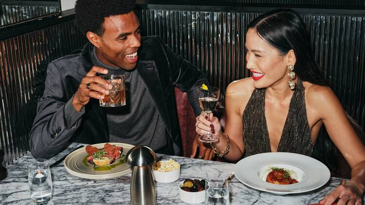 Man and a woman laughing at a restaurant together with plates of food and drinks.