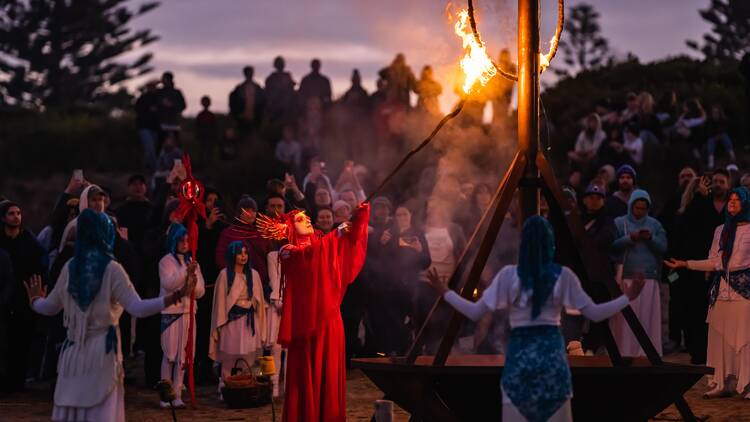 A person in a red cloak lights a wooden installation on fire in front of a crowd.