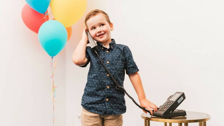 A young boy uses a corded phone in front of some colourful balloons