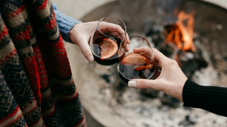 Two people cheers glasses of mulled wine over an open fire.