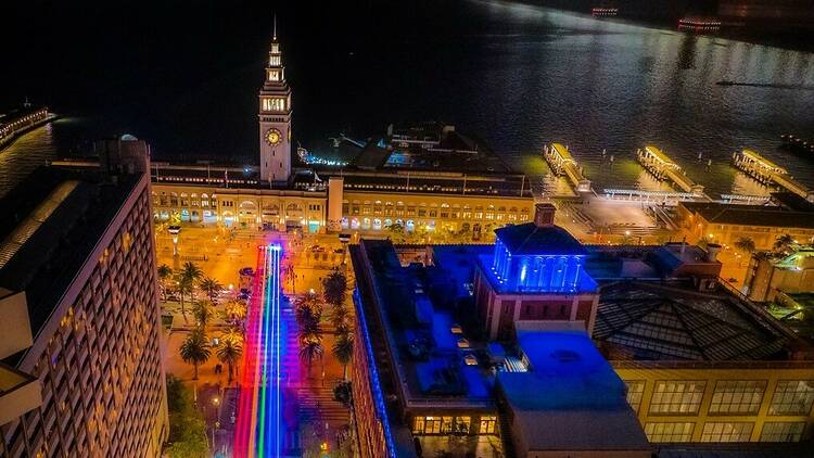 Drone view shows the Ferry Building lit against the dark water of the bay, with the rainbow flag light installation issuing from it down Market Street