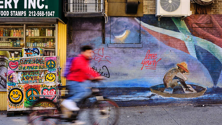 Blurred in action, a young man bikes past a bodega and a mural painted on the wall which shows a man in a boat
