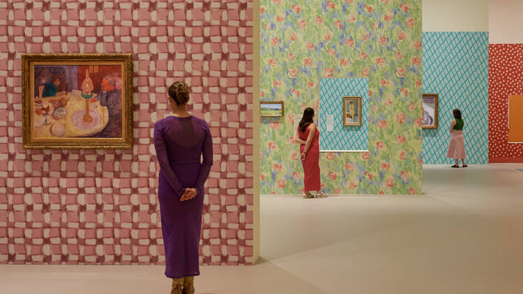 Three women stand in different sections of a gallery looking at colourful artwork.