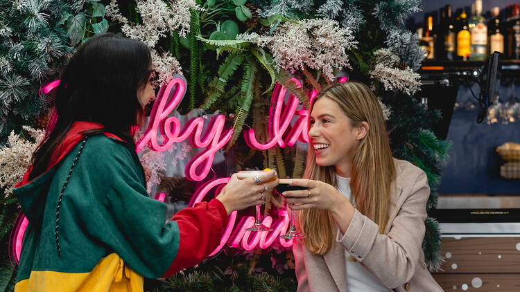 Two female friends clinking cocktails in front of a decorative tree at a bar.