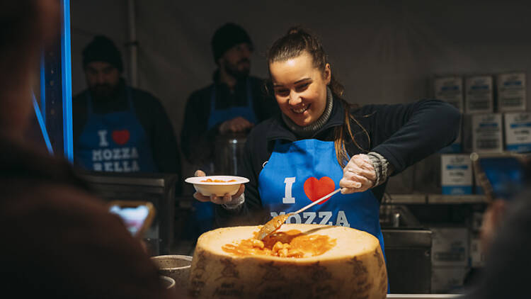Smiling woman doling out pasta from a cheese wheel.