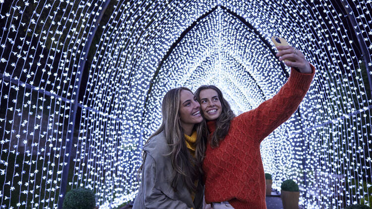 Two women taking a selfie under a fairy-lit tunnel.