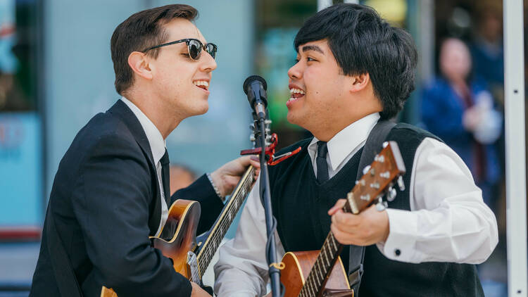 Two men play guitars while standing at a mic.