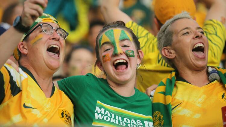 Three women in green and gold cheer on the Matildas