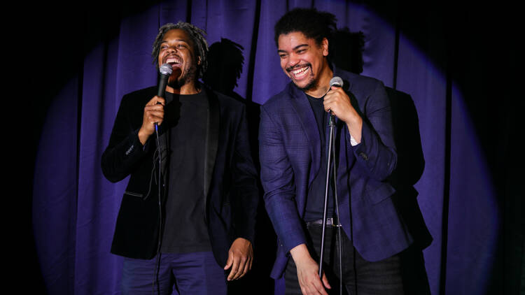 Two comedians on stage in front of a blue curtain.