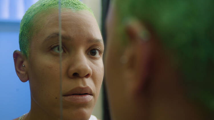 A woman with a green buzzcut stares into a mirror.