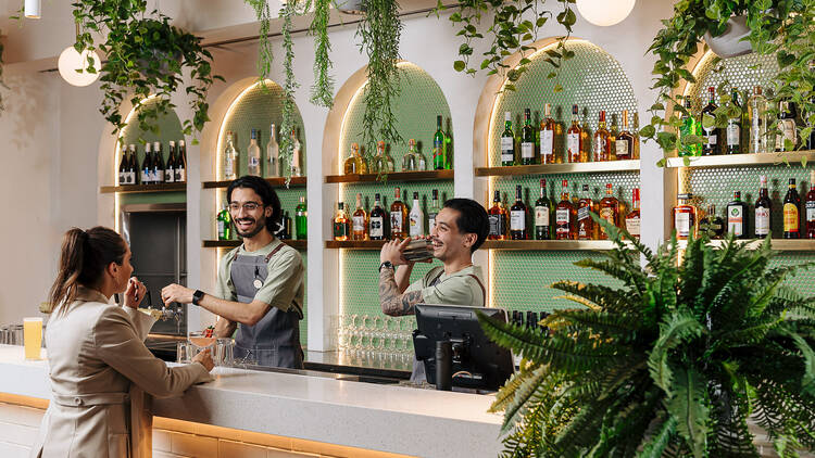 Two bartenders making a cocktail for a woman at a plant-filled bar.