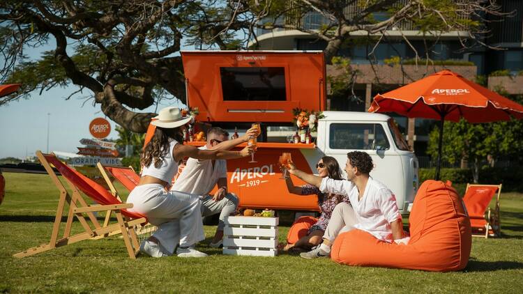 Four people wearing white toast Spritzes in front of the Aperol kombi.