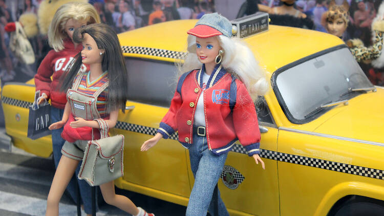 A Barbie doll stands next to a model of a yellow taxi.