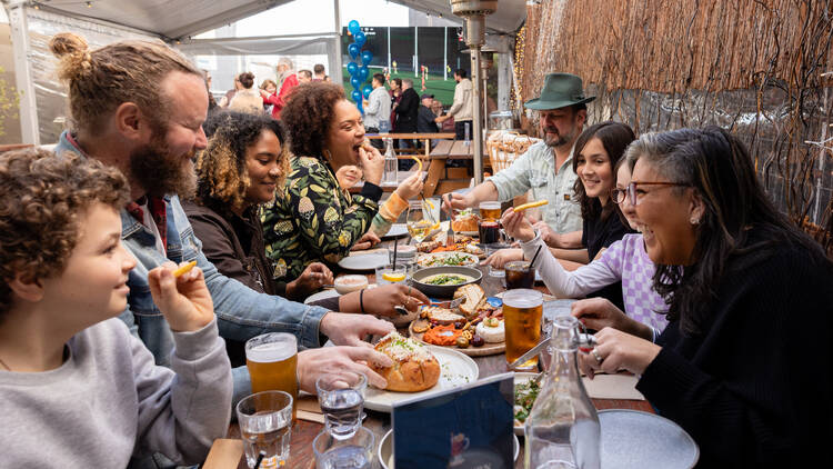 Pub customers laughing and eating around a table of food and pints of beer.