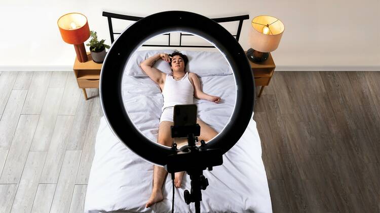 A man in underwear and a singlet lies on a bed, looking up at a phone on a tripod with a ring light.