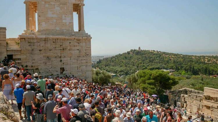 A large crowd of tourists visit the Acropolis of Athens on a hot, sunny day