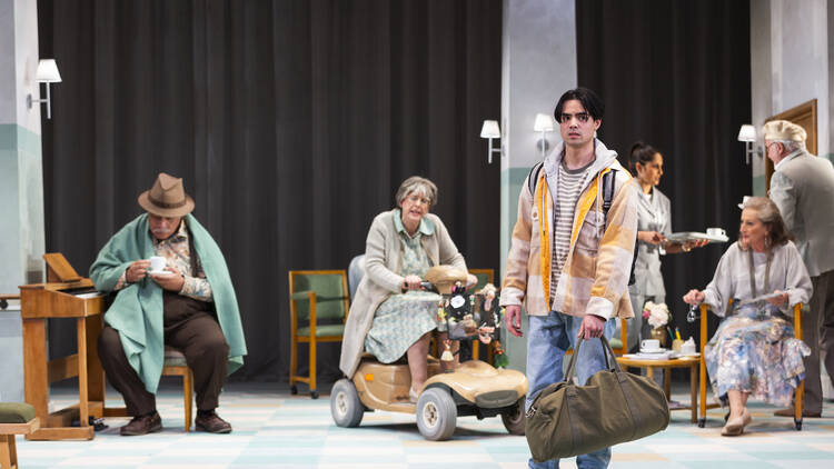 A young man with a duffel bag stands on stage surrounded by elderly actors.