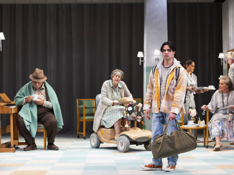 A young man with a duffel bag stands on stage surrounded by elderly actors.