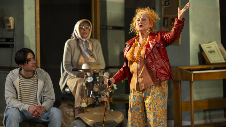 An older woman with orange hair gestures energetically on stage in front of two other actors.