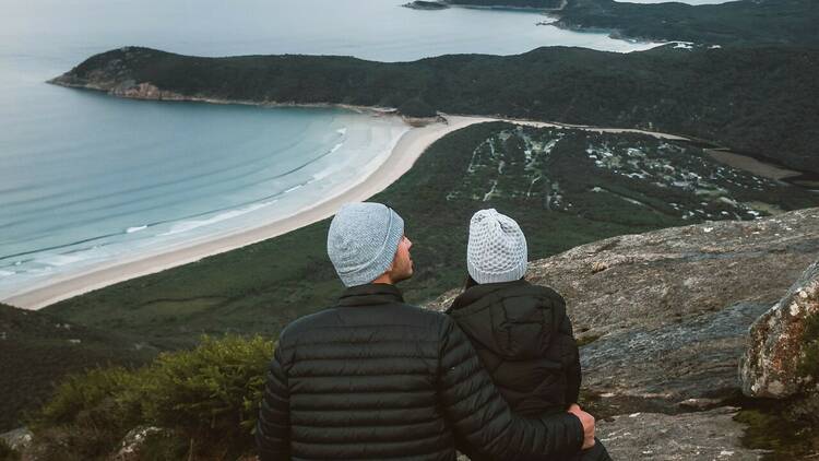 A couple wearing puffer jackets and beanies sits looking out at a coastal vista from above