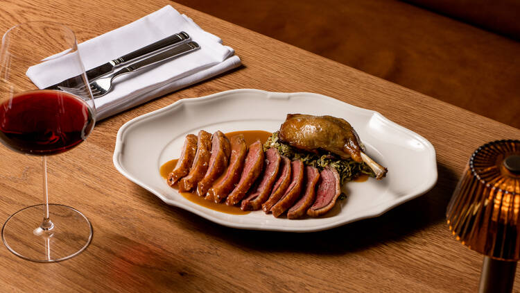 A plate of Great Ocean duck, sugarloaf cabbage and jus on a wooden table with cutlery, a lamp and a glass of red wine.