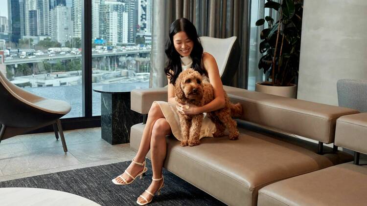 A woman sits on a couch with a dog inside a hotel. 
