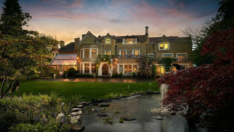 A photograph of a house in the evening with a garden at the bottom of the photo and a pond on the right. 