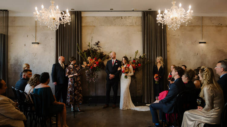 A couple getting married stand in front of an audience inside a function room. 