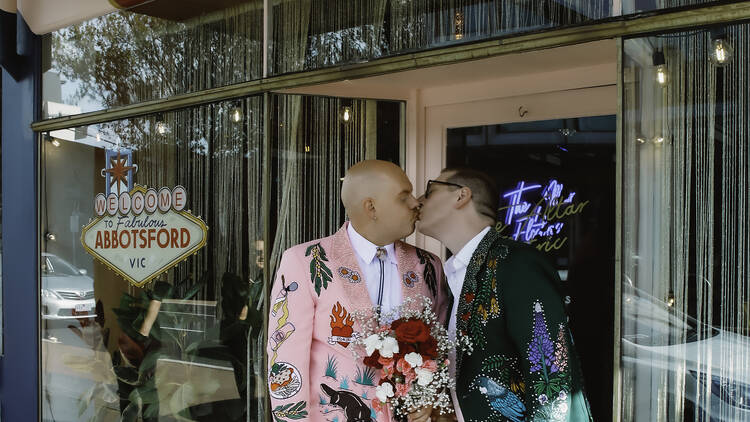 Two men stand outside a wedding chapel, wearing colourful suits and kissing. 