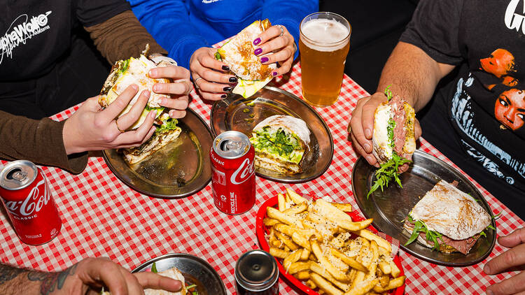 Diners sitting around a table with sandwiches and cans of coke.