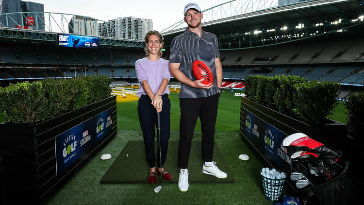 A man and a woman pose with a golf club and a football at Marvel Stadium.
