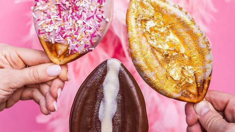 Three vulva-shaped waffles covered in assorted toppings against a pink feathery backdrop.