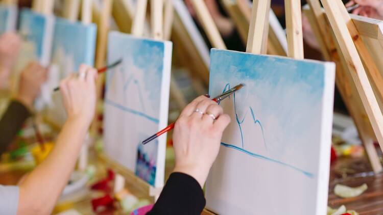 Hands holding paint brushes with blue paint on the canvases. 