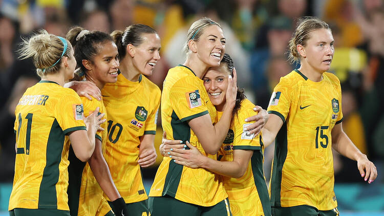 Australian female soccer players wearing yellow and green jerseys celebrate.