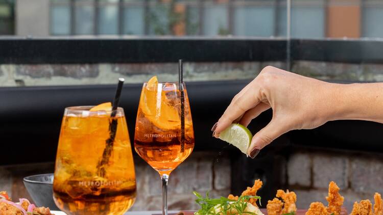 Waiter spritzing a wedge of lemon onto a plate of fried calamari in a rooftop bar setting with Aperol Spritzes.