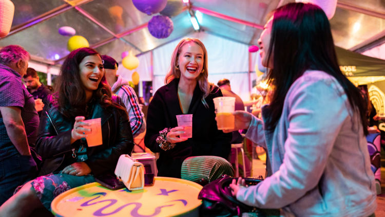 Three girls drinking beers inside a colourful tent