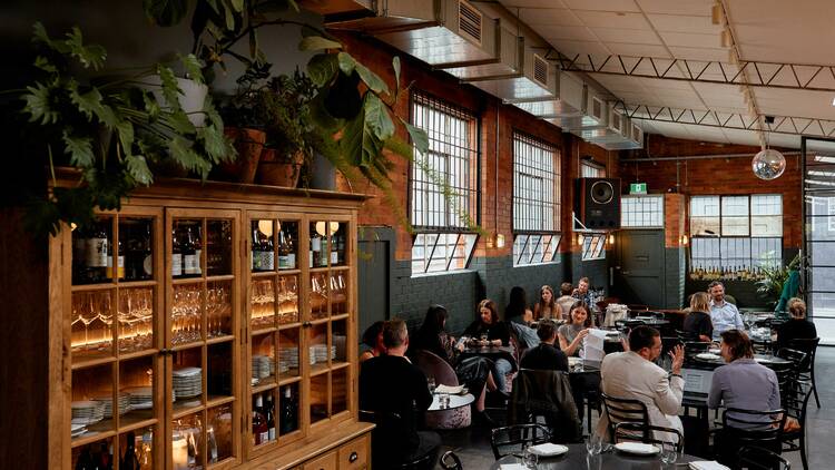 An intimate wine bar featuring wooden cabinets, lush greenery and windows. 