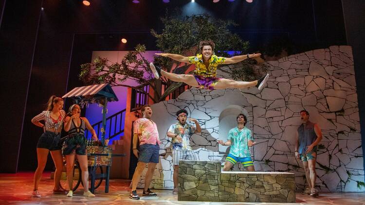 Performers in Mamma Mia! wearing colourful outfits look in awe at one performer doing a leap in the splits.