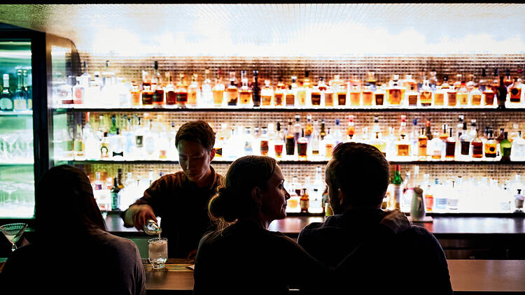 Bartender serving drinks at a backlit, fully stocked bar.