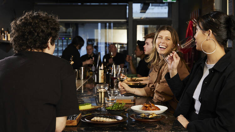 Woman smelling a glass of wine next to a laughing woman at a bar.