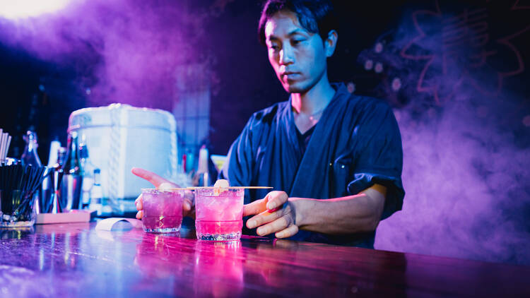 A man standing behind a bar serves up two pink coloured cocktails. 