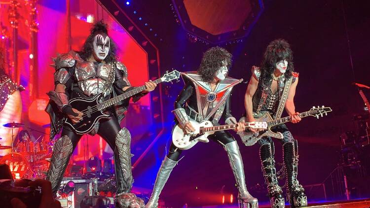 Three members of KISS perform dressed in black costumes and holding guitars.