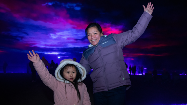 A woman and a child in warm winter clothes raise their arms in front of Borealis on the Lake