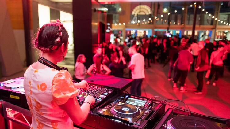 A girl standing behind DJ decks faces out to a crowd of people. 