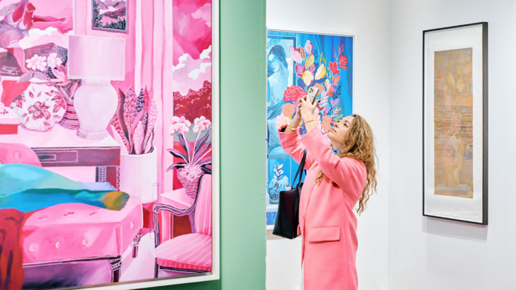 A woman in a pink coat takes a photo of a painting at the Affordable Art Fair.