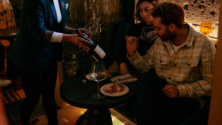 Two people are sitting down and one man is smelling a glass of wine, a third person is holding a bottle.