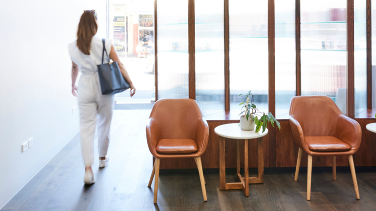 A woman wearing white walks through a modern reception area with tan leather chairs.