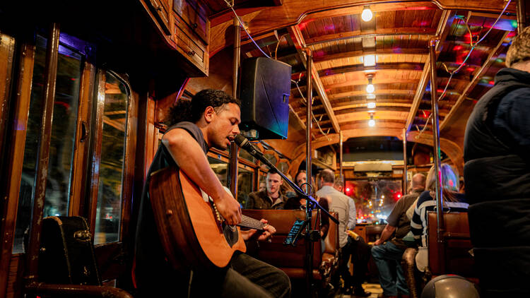 A performer sings into a microphone and plays guitar, inside a tram. 