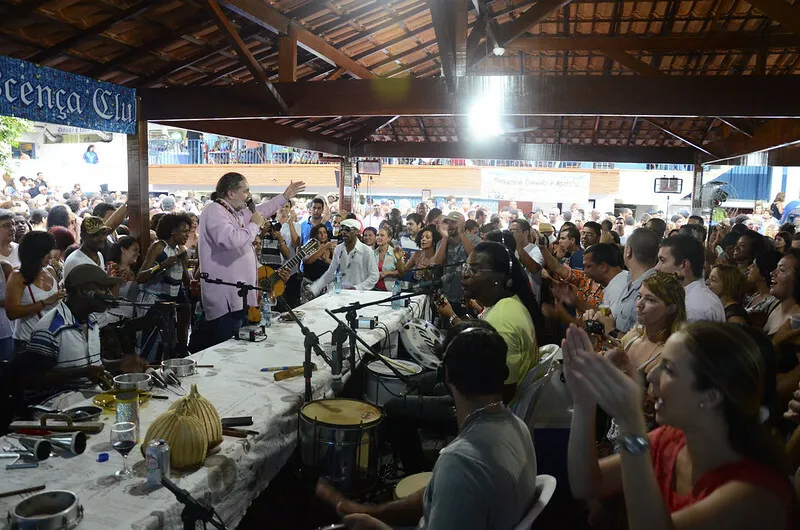Uma roda de samba se apresenta sob um telhado para uma multidão