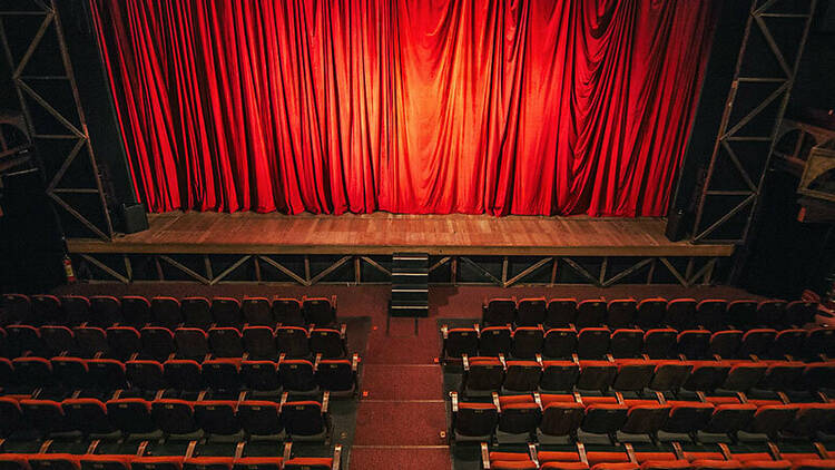 Um palco com cortina vermelha fechada e uma plateia com cadeiras de veludo vermelho